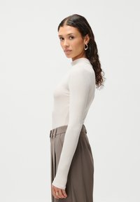 Top de cuello alto de manga larga en beige claro, hecho de un tejido suave. Combinado con pantalones marrones de tiro alto y plisados, que destacan una silueta ajustada.