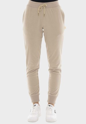 Beige joggerbroek met een elastische tailleband en trekkoord, boorden bij de enkels, zijzakken, gedragen met witte sneakers op een neutrale achtergrond.