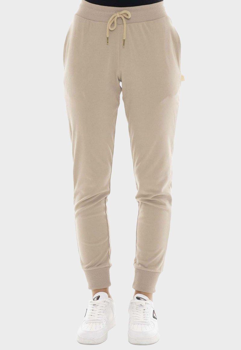 Pantalon de jogging beige avec taille élastique et cordon, ourlets resserrés aux chevilles, poches latérales, porté avec des baskets blanches sur fond neutre.