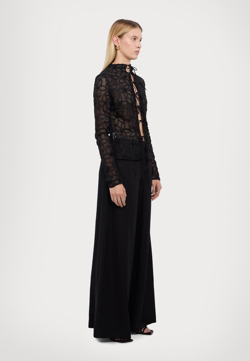 Top court en dentelle noire avec un motif floral, manches longues, et fermeture à nouer ; accompagné de pantalons larges noirs. Des sandales plates complètent le look.