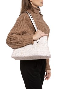 Beige schoudertas met een reliëflogo-patroon, rechthoekige vorm, textielstof en twee contrasterende zwarte handvatten.