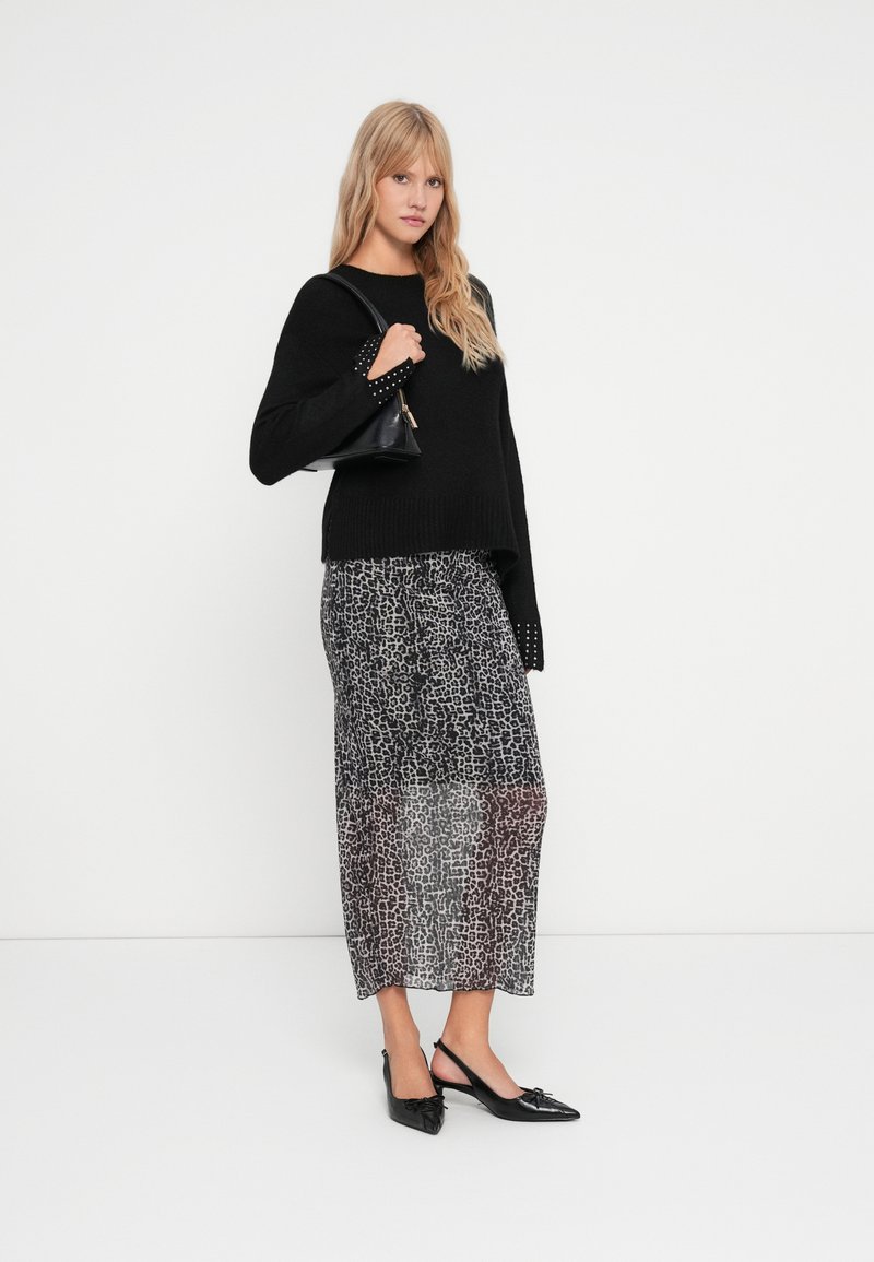 Maglione nero lavorato a maglia con polsini chiodati, abbinato a una maxi gonna trasparente con stampa leopardata in toni di grigio, e scarpe slingback nere.