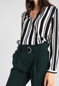 Blouse rayée noir et blanc avec un col en V et des manches longues, associée à un pantalon vert taille haute avec une ceinture à anneau argenté.