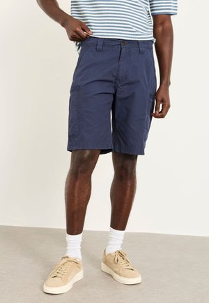 Mann trägt marineblaue Cargo-Shorts, weiße Socken, beige Sneakers und ein blau-weiß gestreiftes Hemd, steht auf einem hellen Boden und Hintergrund.