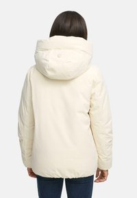 Persoon in een crèmekleurige winterjack met capuchon en donkerblauwe jeans, van achteren te zien tegen een effen achtergrond.