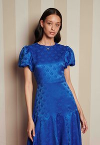 Γυναίκα με μακριά, σκούρα μαλλιά φοράει ένα μπλε φόρεμα με λουλούδια και φουσκωτά μανίκια, στέκεται σε έναν μπεζ ριγέ φόντο.