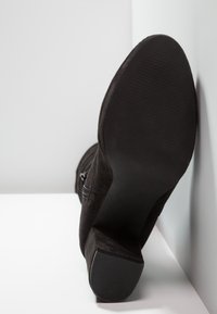 Musta mokkanahkasaapas, jossa on paksu korko, lepäämässä pohja eteenpäin valkoisella pinnalla harmaata seinää vasten.