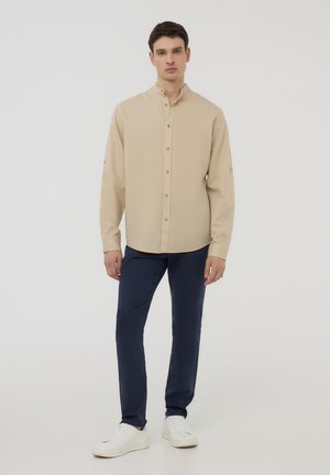 Camicia beige con collo alto, maniche lunghe e bottoni dorati. Abbinata a pantaloni blu scuro e sneakers bianche. Tessuto liscia.