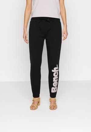 Femme portant un pantalon de jogging noir avec un texte blanc "Bench" sur la jambe gauche et des sandales à talons beiges sur un fond uni.