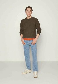 Suéter castanho texturizado por cima de uma camisa laranja, combinado com jeans de ganga azul claro. O modelo usa sapatilhas beje. Design simples, ajuste casual.