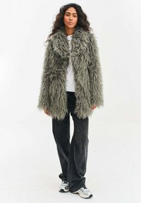 Kobieta z długimi kręconymi włosami, ubrana w szary puszysty futrzany płaszcz, białą koszulę, czarne spodnie o szerokich nogawkach i białe tenisówki, stojąca na białym tle.