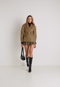 Blazer bege de dupla face com dois bolsos frontais, combinado com um suéter de gola alta escura e uma saia mini. Botas pretas até o joelho e uma bolsa pequena.