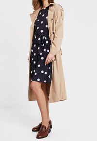 Beige trenchcoat över en marinblå klänning med prickar och knytmidja. Bruna loafers fulländar looken. Slät textur, knälång stil.