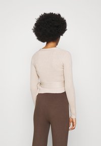 Beżowy sweter w prążki z długimi rękawami, z akcentem związanym w talii. Miękka tekstura i dopasowany krój. Widać również brązowe legginsy w prążki.