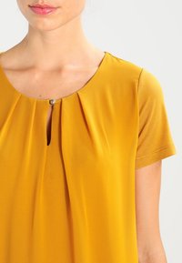 Senfgelbe Bluse mit rundem Ausschnitt, kurzen Ärmeln, einem kleinen Schlüssellochdetail, plissiertem Stoff und einem dekorativen Knopf.