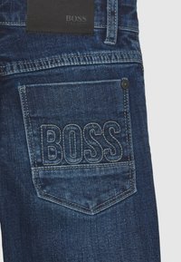 Tmavě modré džíny z denimu s zadní kapsou zdobenou vyraženým logem „BOSS“, zvýrazněným prošíváním a koženou nášivkou nad kapsou.