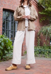 Giacca oversize beige, camicia bianca e pantaloni larghi crema. Scarpe marrone chiaro con suola texturizzata. Ambiente naturale all'aperto con mattoni e vegetazione.