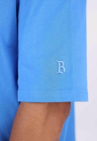 Blå bomull T-skjorte med korte ermer, med en lyseblå brodert "B" på ermet. Glatt tekstur og enkelt design.