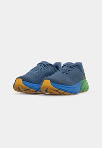 Des chaussures de sport bleues avec une tige texturée, présentant une semelle dégradée en bleu, vert et orange. Les lacets et le logo sont de couleurs contrastées.