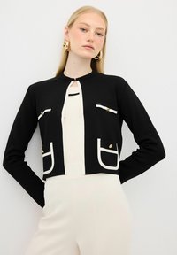 Cardigan cropped nero con accenti bianchi, dotato di scollatura tonda, due tasche frontali e bottoni dorati. Realizzato in materiale a maglia testurizzata.