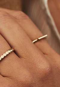 Bagues en or sur une main ; l'une présente des perles rondes, l'autre a un design en barre droite. Peau texturée visible en arrière-plan.