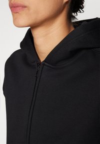 Black zip-up hoodie with a soft texture, featuring a high collar and kangaroo pocket. Design emphasizes simplicity and functionality.