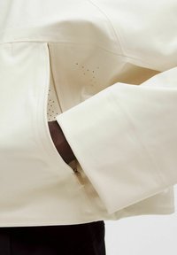 Ljus beige jacka med en zippered ficka, subtila perforeringar, rena linjer och en mjuk, flexibel tygstruktur.