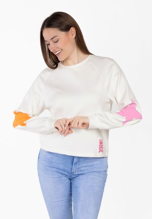 Kobieta uśmiechająca się i patrząca w dół, ubrana w biały sweter z różowo-pomarańczowymi łatami w kształcie gwiazdek na rękawach oraz jasnoniebieskie dżinsy.