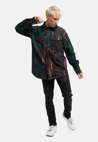 Groene en zwarte gestreepte shirt met twee borstzakken, knoopsluiting en een relaxte pasvorm, gecombineerd met zwarte broeken en witte sneakers.