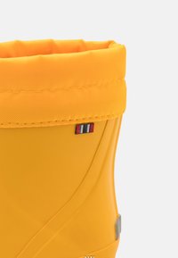 Botte de pluie en caoutchouc jaune avec une tige texturée, un poignet rembourré et un petit emblème tricolore sur le côté avec des accents en métal argenté.