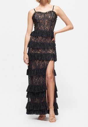 Femme portant une robe maxi noire en dentelle sans manches avec des volants superposés et une fente haute, associée à des talons à brides couleur nude.