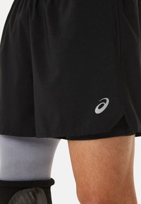 Schwarze Sportshorts aus leichtem Stoff mit einem seitlichen Schlitz und einem reflektierenden Logo am Saum. Grauer Beinschoner teilweise sichtbar.