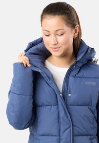 Blå pufferjacka med hög krage och stor huva. Har en framficka med dragkedja och tryckknappar; tyget ser slätt och något glänsande ut.