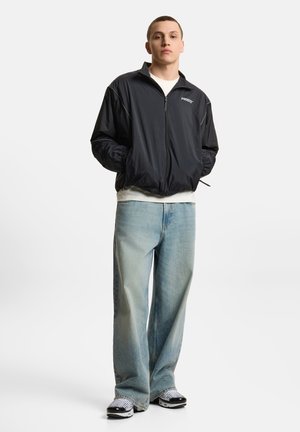 Młody mężczyzna stoi z rękami w kieszeniach, ubrany w czarną kurtkę na zamek, białą koszulę, jasnoniebieskie dżinsy i czarno-białe tenisówki.