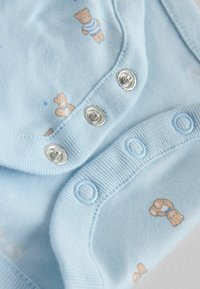 Ljusblå body i mjuk bomull med mönster av björnar i beige. Knäpps med silvriga tryckknappar och ljusblå knappar längs botten.