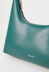 Bolso de mano color verde azulado con una textura suave y cremallera plateada, que presenta un sutil logotipo "Calvin Klein" en blanco en la parte inferior.