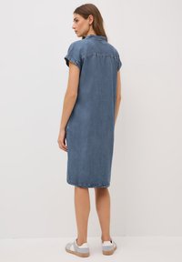 Abito camicia in denim con colletto, maniche rullate corte, design di lunghezza media, colore azzurro chiaro e spacchi laterali. Abbinato a sneakers grigie e bianche.
