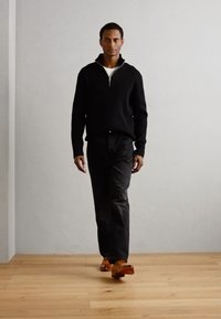 Maglione nero con zip e texture a coste, abbinato a pantaloni scuri e scarpe marroni, indossato da una figura in piedi su un pavimento in legno chiaro.