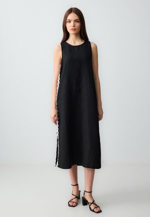 Schwarzes Leinen-Kleid ohne Ärmel mit lockerer Passform, das einen kontrastierenden gewellten weißen Seitenbesatz und ein knöchellanges Design aufweist. Schwarze Riemchen-Absätze werden getragen.