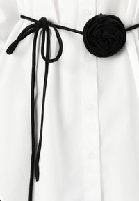Black ribbon belt with a large fabric rose detail, worn over a white shirt featuring button closures and a smooth texture.