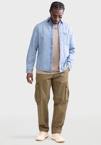 Hellblaues Denim-Button-Up-Hemd über einem beigen T-Shirt, kombiniert mit khakifarbenen Cargo-Hosen und weißen Sneakers. Merkmale sind Taschen an den Hosen.