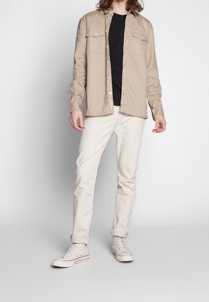 Camicia beige con bottoni e due tasche sul petto, indossata sopra una t-shirt nera, abbinata a jeans bianchi aderenti con fondo stretto e sneakers chiare.