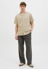 Camicia beige a maniche corte con un motivo geometrico, tessuto leggero, chiusura con bottoni davanti, abbinata a pantaloni grigio scuro e scarpe beige.