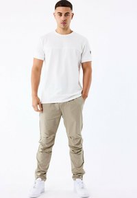 Camiseta blanca de manga corta con un panel texturizado, combinada con pantalones cargo de color beige claro y zapatillas blancas, de pie contra un fondo liso.