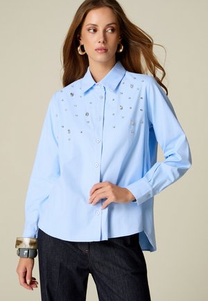 Vrouw draagt een lichtblauw overhemd met knopen en strassdetails op de borst en een donkere broek, gecombineerd met grote oorringen en armbanden.