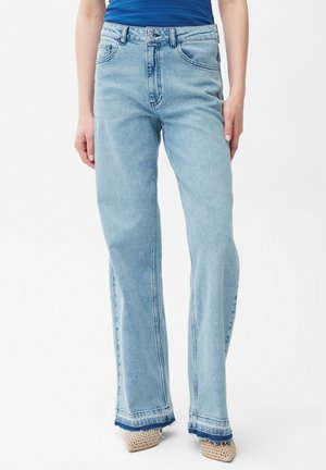 Osoba nosząca jasnoniebieskie, proste jeansy z wysokim stanem i surowymi brzegami, w połączeniu z beżowymi, plecionymi, szpiczastymi butami oraz niebieską bluzką.