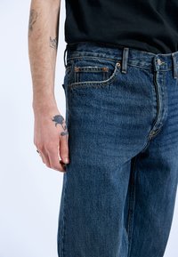 Jeans de mezclilla azul oscuro con corte relajado, diseño de cinco bolsillos, costuras de cobre y textura visible. Mano con un tatuaje descansando de lado.