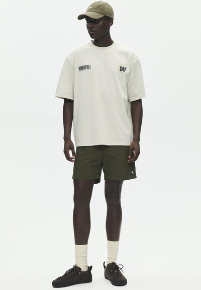 Άνδρας μοντέλο φοράει μπλούζα σε μπεζ oversized, χακί σορτς, μπεζ κάλτσες, μαύρα αθλητικά παπούτσια και καπέλο μπέιζμπολ σε χακί, στέκεται μπροστά σε λευκό φόντο.