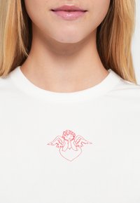 Λευκό βαμβακερό μπλουζάκι με κόκκινο γραφικό αγγέλου με φτερά, που κρατά τα χέρια του στο πρόσωπο, μέσα σε σχήμα καρδιάς.