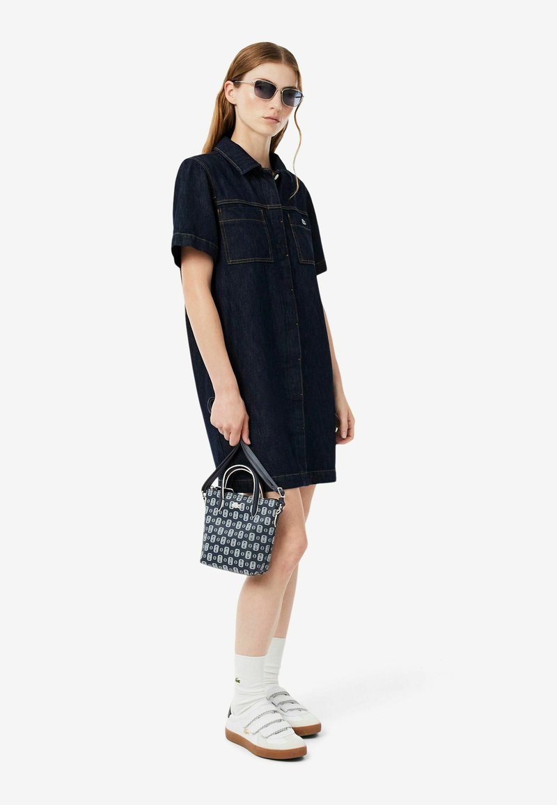 Frau trägt ein dunkles Denim-Hemdkleid, weiße Socken, weiße Sneakers, Sonnenbrille und hält eine gemusterte Handtasche mit schwarzen Griffen.